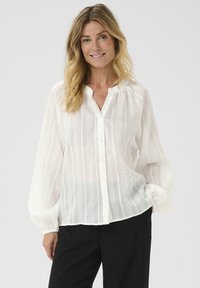 Camisa blanca ligera y de corte relajado, con rayas verticales y mangas largas abullonadas, combinada con pantalones negros.