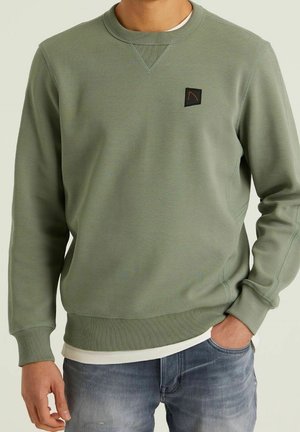 Hombre con sudadera de cuello redondo verde oliva con puños y dobladillo acanalados, pequeño logo de parche negro en el pecho, combinada con vaqueros azules.