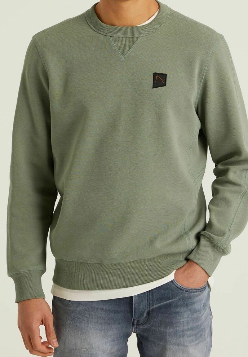 Homme portant un sweat-shirt à col rond vert olive avec poignets et ourlet côtelés, petit logo patch noir sur la poitrine, associé à un jean bleu.