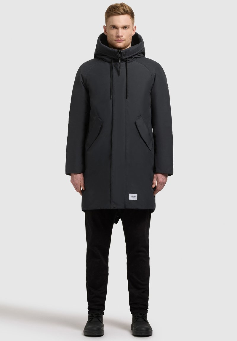 Juodas parka paltas su kapišonu, priekiniu užtrauktuku ir stygomis. Yra šoninės kišenės ir logotipo aplikacija ant apvado. Dėvimas su juodomis kelnėmis ir batais.
