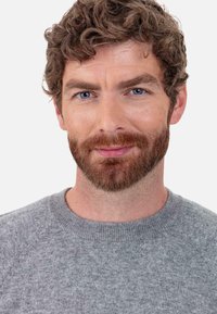 Porträt eines Mannes mit lockigem braunem Haar und vollem Bart, der einen hellgrauen Strickpullover mit rundem Ausschnitt und strukturiertem Stoff trägt.