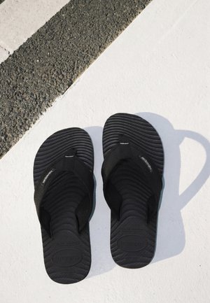 Coppia di infradito nere Havaianas posizionate su una superficie bianca accanto ad asfalto testurizzato con una linea bianca.