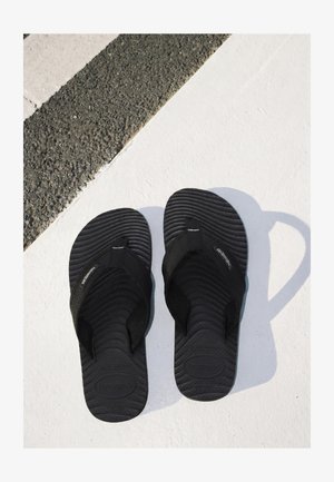 Coppia di infradito nere Havaianas posizionate su una superficie bianca accanto ad asfalto testurizzato con una linea bianca.
