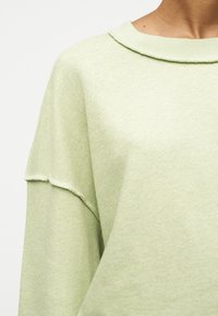 Nærbilde av lys grønn genser med senket skuldersøm og rå kant på kragen, båret av en person mot hvit bakgrunn.