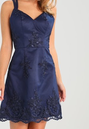 Femme portant une robe sans manches bleu marine avec une broderie en dentelle florale et un ourlet festonné sur un fond clair uni.
