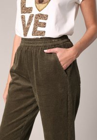 Olijfgroene corduroy broek met een ribbelstructuur, elastische tailleband en zijzakken. Gecombineerd met een witte T-shirt met een grafische opdruk en teksturele lettertype.