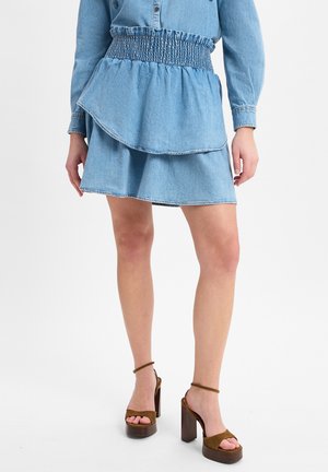 Jupe en denim avec taille smockée, design en couches et ourlet effiloché. Accompagnée de sandales à talons hauts marron avec une large bride.