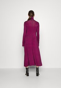 Long, fitted dress in bright pink with a textured pattern. Features a high collar and long sleeves. Accompanied by black, shiny ankle boots.