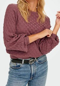 Bordeauxfarbenes, strukturiertes Sweatshirt mit einem gewellten Muster, gerippten Bündchen und einer lockeren Passform. Kombiniert mit blauen, hochgeschnittenen Jeans und einem Gürtel mit Doppelschließe.