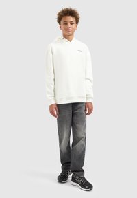 Tiener met krullend haar die een witte hoodie, vervaagde zwarte jeans en zwarte sportsneakers draagt, staand tegen een effen achtergrond.