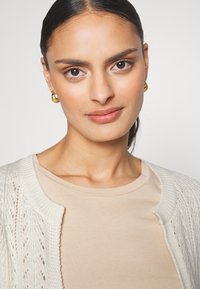 Beige t-shirt paired with a cream knitted cardigan featuring a lace pattern. Gold hoop earrings accent the look. Smooth skin and neutral makeup.