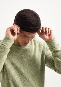 Bruine gebreide beanie op een persoon die het aanpast, gecombineerd met een lichtgroene textuurtrui, die ribpatronen en zachte stof toont.