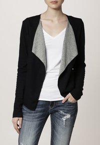 Svart cardigan med en ljusgrå draperad krage, mjukt tyg, öppen frontdesign och ribbade ärmar. Bärs över en vit V-ringad t-shirt.