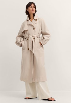 Woman wearing a beige belted trench coat with puffed sleeves, wide-leg white pants, and black sandals, standing against a plain background.