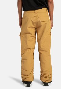 Beżowe spodnie narciarskie o luźnym kroju, z kieszeniami cargo po bokach, wzmocnionymi szwami i elastycznymi mankietami na kostkach.