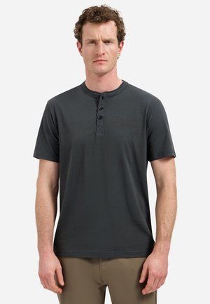 Mann mit kurzen lockigen Haaren, der ein dunkelgraues Henley-Shirt mit kurzen Ärmeln und hellbraune Hosen trägt, vor einem schlichten Hintergrund.