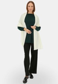 Biały szal zarzucony na ciemnozielony sweter, w połączeniu z czarnymi spodniami. Model ma na sobie czarne buty, co podkreśla smukłą sylwetkę.