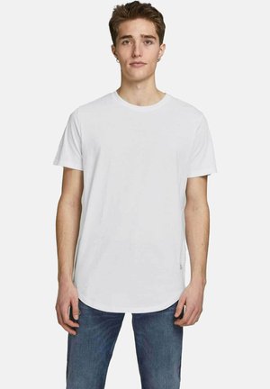 Jeune homme aux cheveux bruns courts portant un t-shirt blanc uni à manches courtes et un jean bleu, debout devant un fond clair.