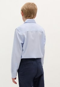 Camicia azzurra chiara con maniche lunghe e chiusura con bottoni, dotata di yoke posteriore e dettaglio con pieghe. Abbinata a pantaloni blu scuro, con tasche posteriori.