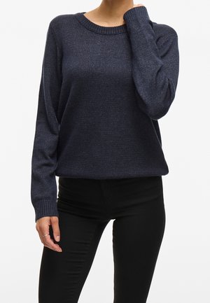 Femme portant un pull en maille bleu foncé à manches longues et un pantalon noir, posant avec une main touchant son cou.