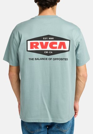 Camiseta de algodón turquesa con un gran logotipo de RVCA en negro y rojo en la parte posterior, con el texto "EL BALANCE DE LOS OPUESTOS." Mangas cortas.