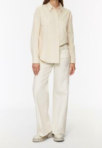 Camisa beige de manga larga con botones en la parte delantera, combinada con pantalones de pierna ancha color crema y botas beige gruesas. Textura suave, diseño minimalista.