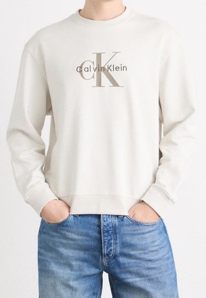 Osoba nosząca kremową bluzę Calvin Klein z logo na piersi, w połączeniu z niebieskimi dżinsami, ręce wsunięte w kieszenie.