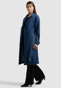 Kvinde iført en lang blå denim dobbeltspændt frakke, sorte bukser og sorte hælede sko, stående mod en ensartet lys baggrund.