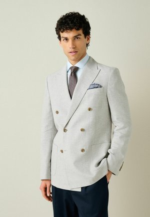 Man wearing light gray double-breasted blazer with brown buttons, blue shirt, patterned tie, and navy pants, posing against beige background.