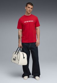 Rød bomulds t-shirt med hvidt logo, løse denim jeans og en stor hvid gymnastiktaske med sorte detaljer og logo. Glatte teksturer hele vejen igennem.