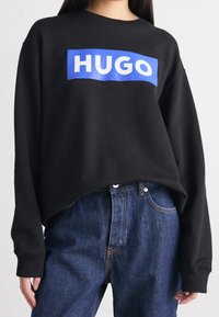 Black sweatshirt with a blue rectangular logo reading "HUGO," featuring long sleeves and a relaxed fit, paired with blue jeans.