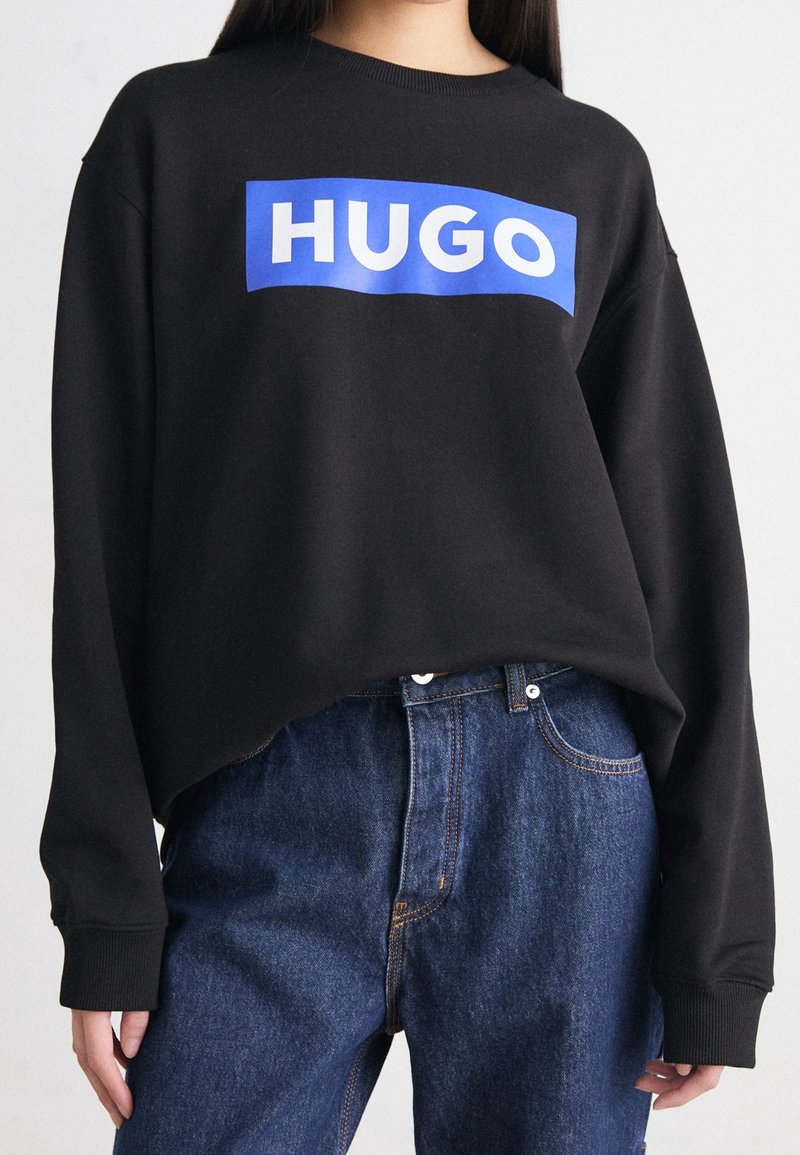 Black sweatshirt with a blue rectangular logo reading "HUGO," featuring long sleeves and a relaxed fit, paired with blue jeans.