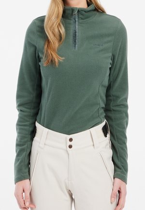 Frau trägt einen grünen Fleecepullover mit Viertelreißverschluss und weißen, hoch taillierten Hosen mit Fronttaschen.