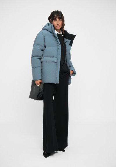 Padded modra puffer jakna s kapuco, z dvema sprednjima žepoma, nosi se čez črni pulover in široke črne hlače. Vključena je črna torbica.