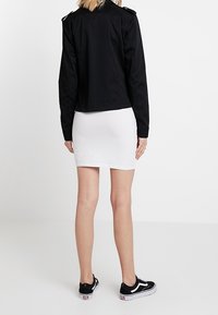 Osoba stojící zády, oblečená v černé bundě, bílé mini sukni a černo-bílých teniskách, na jednobarevném bílém pozadí.