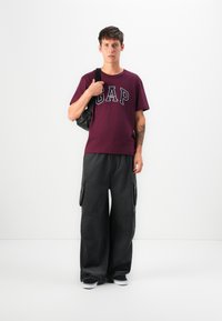 Ein junger Mann trägt ein burgunderfarbenes GAP-T-Shirt, lockere schwarze Cargo-Hosen, schwarze Sneakers und hält eine schwarze Umhängetasche. Er steht vor einem weißen Hintergrund.
