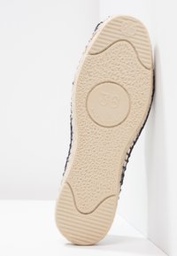 Gummisohle eines Schuhs mit einer strukturierten beigen Oberfläche, ausgestattet mit einem runden Logo und einem niedrigen Schnitt, umrandet von einem dunkelblauen Obermaterial.