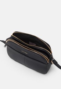 Czarna skórzana torba na ramię z dwoma zamykanymi na suwak przegródkami, złotymi akcentami metalowymi i wytłoczonym logo marki z przodu. Gładka faktura.