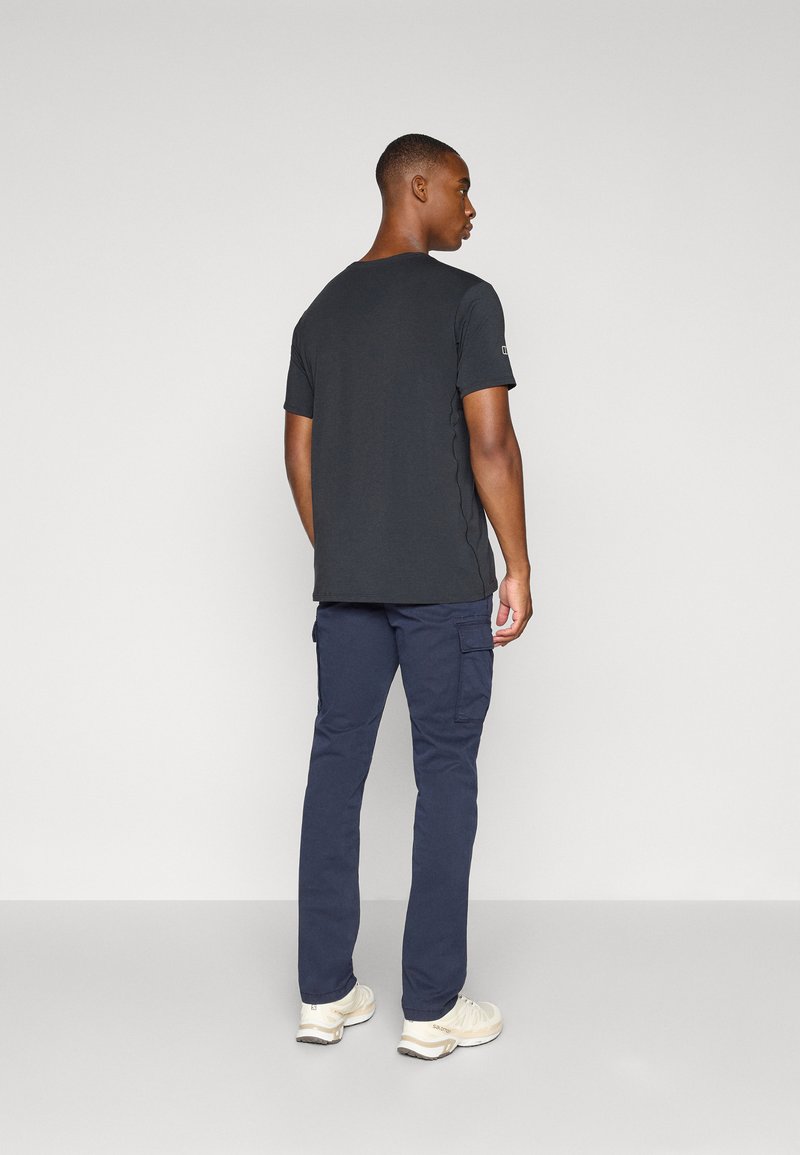 T-shirt noir à manches courtes avec un col rond, porté avec un pantalon cargo bleu marine et des baskets beiges. Le t-shirt présente des coutures latérales et un logo sur la manche.