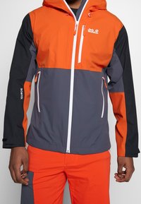 Waterdichte jas in oranje en grijs, met een capuchon, witte rits en logo. Contrasterende panelen met een gladde textuur en verstelbare mouwen.