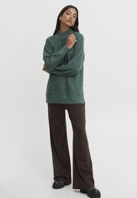 Grüner gerippter Strickpullover mit hohem Kragen und langen Ärmeln, kombiniert mit braunen weiten Hosen und schwarzen Plateaustiefeln.