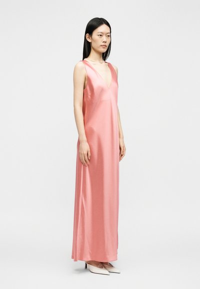 Γυναίκα με ίσια μαύρα μαλλιά, που φοράει ένα ροζ σατέν μακρύ φόρεμα χωρίς μανίκια και λευκά μυτερά τακούνια, στέκεται μπροστά σε ένα απλό φωτεινό φόντο.