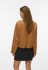 Žena s dlouhými rovnými vlasy, oblečená v hnědé semišové bundě a černé sukni, stojí čelem k bílému pozadí.