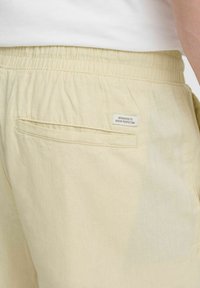 Lys gule shorts med elastisk midje, med en baklomme og en liten, sydd etikett som sier "Dedikert til denim-perfeksjon."