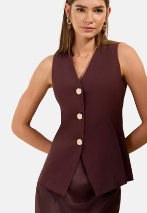 Woman wearing sleeveless dark burgundy vest with three large gold buttons and matching skirt, standing with hands behind back.