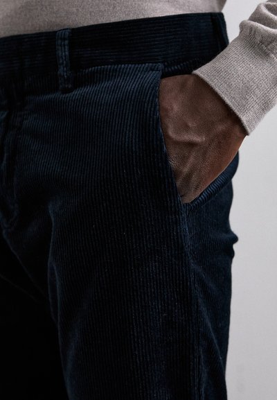 Navy corduroy pants with a textured ribbed pattern, featuring a side pocket and a close-up of a hand resting in the pocket.