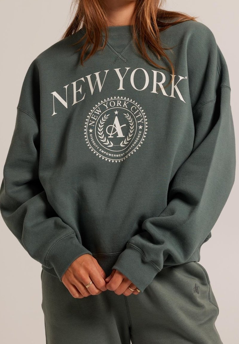 Tumman vihreä huppari, jossa on "NEW YORK" ja pyöreä graafinen logo. Varustettu ribbikämmensuilla ja rento leikkaus. Pehmeä, teksturoitu kangas.