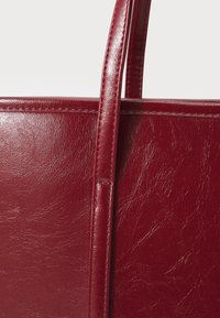 Sac fourre-tout en simili cuir rouge avec une texture lisse et brillante, doté de longues poignées et d'une forme rectangulaire. Coutures visibles accentuant les bords.