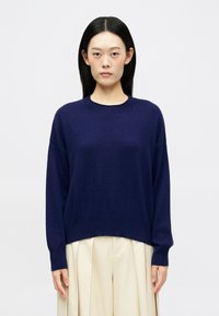 Pull en maille bleu marine avec un col rond, coupe décontractée et poignets côtelés. Assorti à un pantalon plissé en crème.