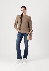 Frau mit geflochtenem Haar, die einen braunen Strickpullover, blaue Jeans, weiße Sneakers trägt und eine beigefarbene Handtasche vor einem schlichten weißen Hintergrund hält.